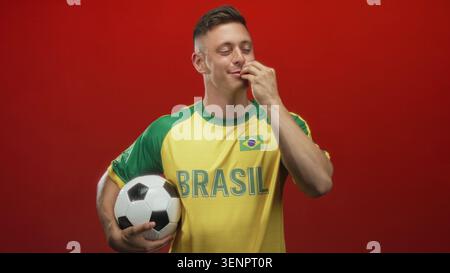 Homme tient le ballon de football sous le bras et retourne les doigts en studio tout en portant le maillot brésilien et en souriant avec les yeux fermés ; célébration de fierté. Banque D'Images