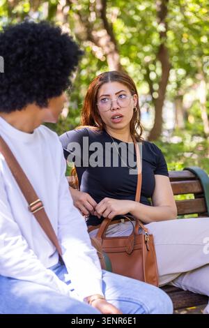 Amis adultes assis sur le banc en bois dans le parc, femme en haut noir tenant sac à main, homme écoutant Banque D'Images