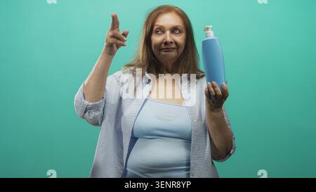 Femme senior tient une bouteille de savon bleue tout en pointant l'index vers la tempe dans le studio ; démonstration de produit réfléchie. Banque D'Images