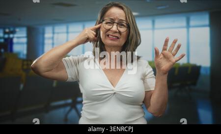 Une femme hispanique âgée pointe du doigt vers la tempe avec la paume ouverte soulevée au milieu des sièges de départ de l'aéroport ; attention réfléchie. Banque D'Images