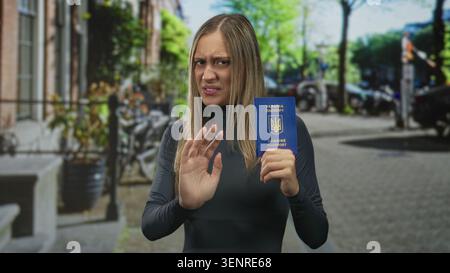 Femme tenant un passeport bleu, paume levée dans une rue urbaine animée avec des voitures et des vélos garés ; réticence. Banque D'Images