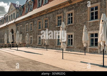 Erfurt, Allemagne - 27 mars 2026. Façade de bâtiment historique en pierre avec toit en tuiles rouges, fenêtres, terrasse en bois, parasols fermés, lumière du soleil, et long sh Banque D'Images