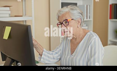 Femme senior pointe l'index sur le moniteur d'ordinateur, main visible, portant des lunettes dans un bureau de bâtiment ; indépendance de concentration. Banque D'Images