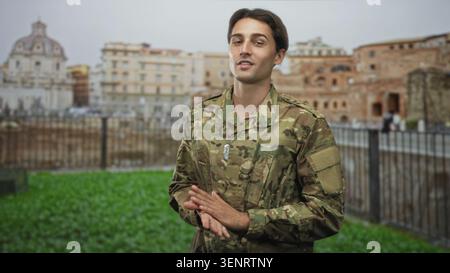 Homme soldat en uniforme de camouflage avec des étiquettes de chien, mains attachées devant, debout près d'un bâtiment historique et une clôture de fer ; devoir de fierté. Banque D'Images