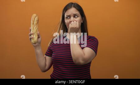 Femme tenant une baguette mord des ongles en studio avec fond orange et chemise rayée ; anxiété malaise hésitation. Banque D'Images