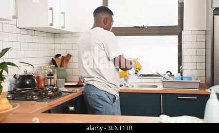 Homme lavant la vaisselle dans la cuisine moderne de la maison portant des gants jaunes et des vêtements décontractés Banque D'Images