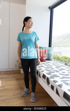 Femme debout regardant par la grande fenêtre dans la chambre portant un haut et des leggings sarcelle, regarder les oreillers de banc Banque D'Images