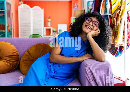 Jeune femme hispanique avec des cheveux bouclés et des lunettes reposant sur un canapé violet à la maison, souriant brillamment et expérimentant le bonheur dans un cadre vibrant et coloré Banque D'Images