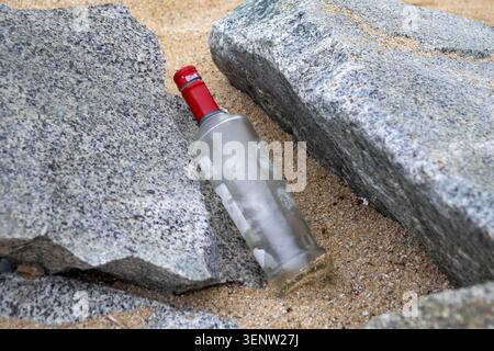 Bouteille de verre abandonnée logée parmi de grands rochers côtiers sur une plage de sable. Image conceptuelle représentant les déchets, la pollution marine, néglec environnemental Banque D'Images