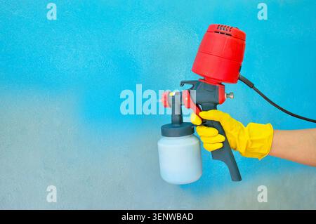 La main d'un ouvrier dans un gant jaune tient un pistolet de pulvérisation à main avec un compresseur en tête. Peindre le mur de la maison en bleu. Ménage Banque D'Images