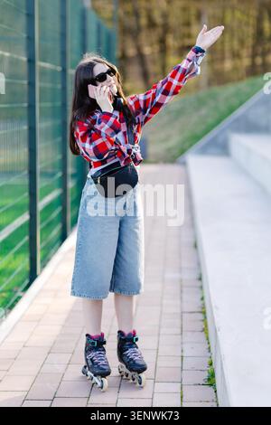 Jeune femme patinant à roulettes dans le parc, utilisant son téléphone tout en saluant un taxi, profitant d'une journée ensoleillée remplie de liberté et de plaisir Banque D'Images