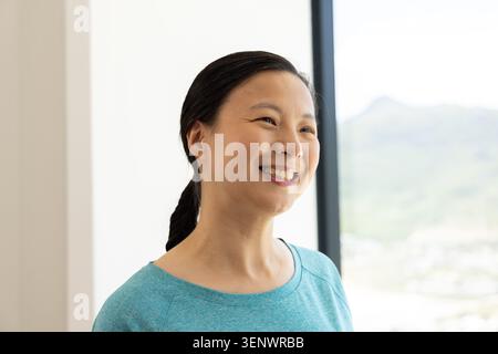Femme mature asiatique souriante, debout dans une pièce lumineuse près d'une grande fenêtre à cadre sombre portant un haut sarcelle Banque D'Images