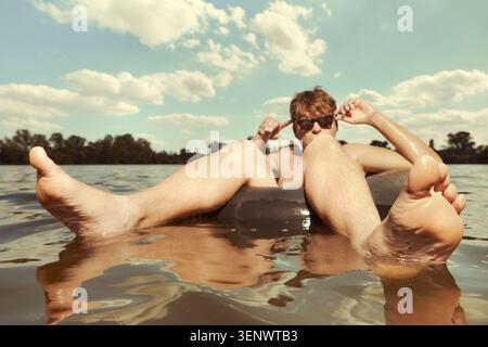 Homme drôle dans des lunettes de soleil nageant dans le lac sur la chambre à air Banque D'Images
