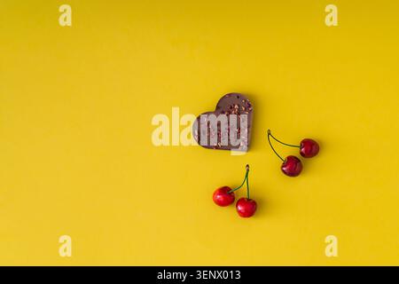 Chocolat en forme de coeur et cerises rouges fraîches sur fond jaune avec espace de copie. Concept de cuisine romantique. Pose à plat. Bannière vue de dessus. Banque D'Images