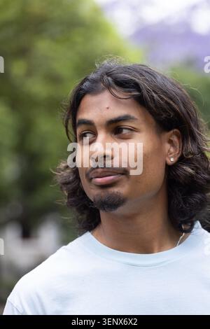 Homme indien dans la vingtaine debout dans le parc portant T-shirt bleu clair, boucle d'oreille clous, collier Banque D'Images