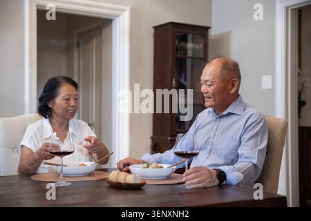 Couple asiatique, femme senior et homme d'âge moyen, partageant repas et vin rouge à la table à manger en bois Banque D'Images