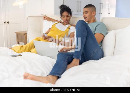 Couple diversifié naviguant dans le lit, femme en salopette jaune tenant la carte de crédit à l'aide d'un ordinateur portable Banque D'Images
