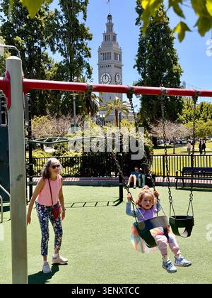 Grand-mère poussant sa petite-fille sur une balançoire dans un parc à San Francisco, Californie. Tour de l'horloge de 245 mètres au sommet de l'historique Ferry Building, à l'arrière. Banque D'Images