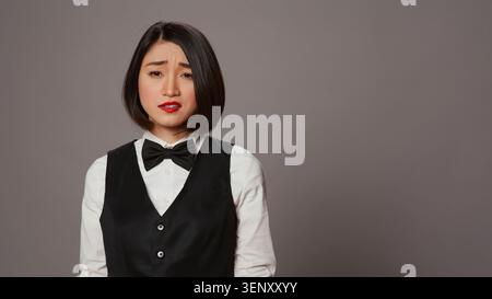Réceptionniste ennuyé étant confus et surchargé de travail en studio, posant avec confusion et fatigue sur fond gris, administrateur asiatique se sentant incertain et incertain de quelque chose. Banque D'Images