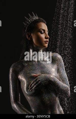 Une femme sereine avec les yeux fermés, son corps couvert de paillettes holographiques chatoyantes, porte une couronne royale. Portrait studio sur fond noir avec dra Banque D'Images