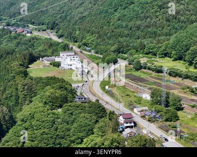 Des collines verdoyantes entourent une route rurale sinueuse. Des bâtiments blancs parsèment le paysage près du bord de la route. Les voitures se déplacent lentement le long d'un chemin d'asphalte courbe. Arbres et f Banque D'Images
