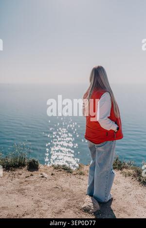 Femme falaise océan. Jeune femme debout sur un bord de falaise surplombant la mer bleue calme sous un ciel clair avec espace de copie Banque D'Images
