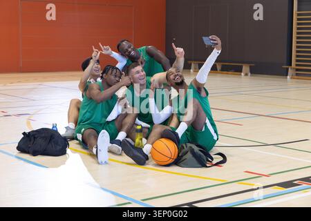 Équipe masculine diversifiée en maillots verts sur le terrain de gymnastique prenant selfie avec téléphone basket-ball et équipement Banque D'Images