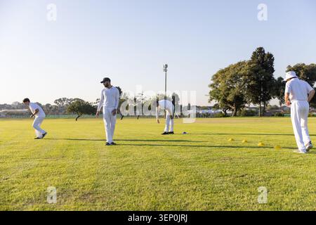 Équipe masculine diversifiée pratiquant des exercices sur terrain de cricket dans des kits blancs et des cônes bleus jaunes Banque D'Images