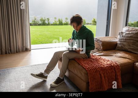 Homme asiatique assis sur un canapé en cuir marron à l'aide d'une tablette avec stylet dans un salon moderne Banque D'Images