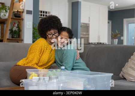 Mère et fille câlins sur un canapé gris à la maison portant un pull jaune moutarde avec des bacs transparents Banque D'Images