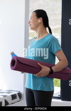 Femme afro-américaine mature tenant un tapis de yoga violet et une bouteille d'eau par fenêtre, portant des écouteurs Banque D'Images
