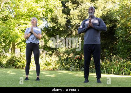 Couple diversifié dans l'usure athlétique debout avec les mains pressant à la poitrine, méditant sur la pelouse de jardin Banque D'Images