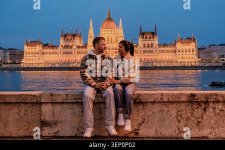 Budapest, Hongrie présente une belle scène nocturne le long du Danube. Un couple se détend sur la rive, en admirant le magnifique bâtiment illuminé du Parlement en arrière-plan. Banque D'Images