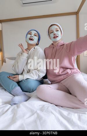 Deux amies assises sur le lit portant des masques faciaux bandeaux en éponge pastel loungewear dans la chambre Banque D'Images