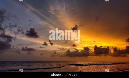 Coucher de soleil tropical doré sur l'océan avec des nuages spectaculaires, un horizon lumineux et de douces vagues reflétant la lumière chaude du soir dans un paysage marin serein. Banque D'Images