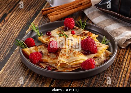Crêpes dorées surmontées de framboises fraîches et de brins de romarin sur une assiette grise, posées sur une table en bois rustique avec des bâtons de cannelle et un napki rayé Banque D'Images