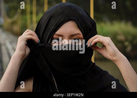 Femme aux yeux bleus frappante regardant sous un niqab noir, visage et tête partiellement couverts, portrait serein et contemplatif véhiculant une identité culturelle Banque D'Images
