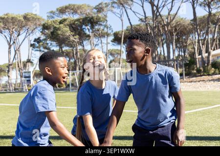 Des enfants divers se tenant la main riant sur un terrain de football gazonné portant des T-shirts bleus près du filet de but Banque D'Images