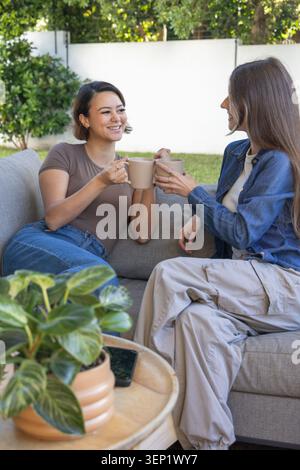 Diverses amies féminines assis et clinking tasses beige sur le canapé gris clair sur le patio arrière Banque D'Images
