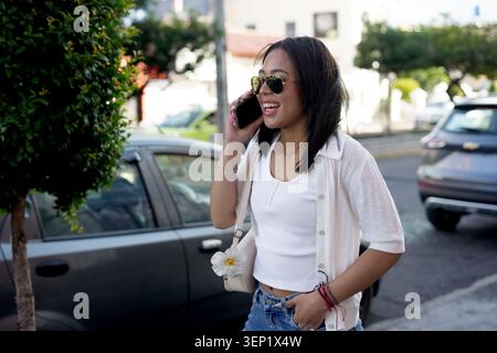Jeune femme portant des lunettes de soleil et un haut blanc, parlant joyeusement sur son smartphone tout en se tenant dans une rue de la ville, communiquant et profitant d'un cheerf Banque D'Images