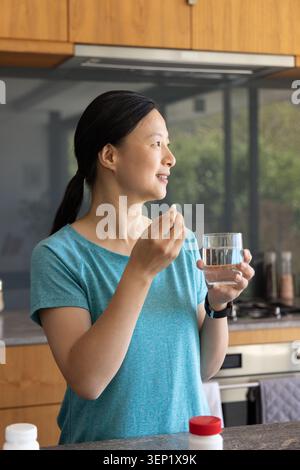 Femme asiatique mature portant chemise sarcelle tenant la pilule et le verre d'eau dans la cuisine avec bouteille de bouchon rouge Banque D'Images