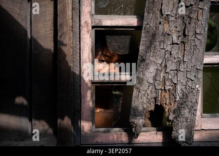 Oeil de femme regardant à travers une fenêtre cassée d'une vieille maison en bois abandonnée, représentant des concepts de pauvreté, de difficultés et de lutte dans un en négligé Banque D'Images