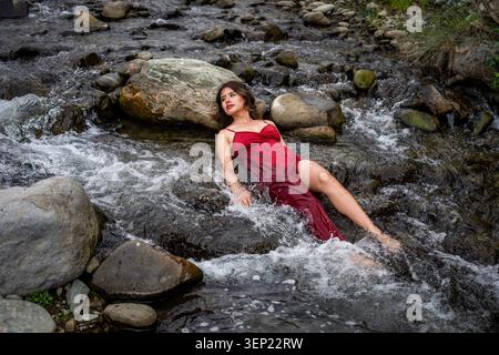 Sereine femme aux cheveux foncés dans une robe rouge couchée dans une rivière rocheuse claire, les yeux fermés, les lèvres rouges et les cheveux longs coulant dans l'eau fraîche, harmonie paisible avec Banque D'Images