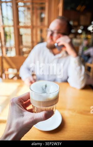 POV de main tenant latte dans un café avec homme flou au téléphone en arrière-plan, lumière du soleil chaude, faible profondeur de champ, concept de distraction, travail à distance, communication et style de vie moderne Banque D'Images