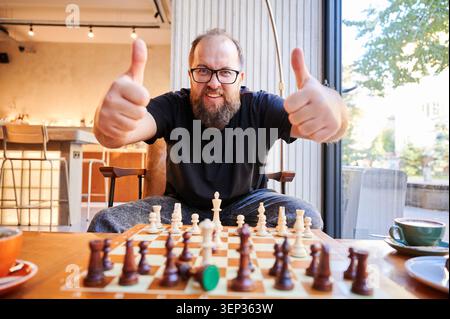 Homme joyeux célébrant la victoire. Homme heureux abandonnant les pouces au café. L'homme à la barbe partage un geste joyeux de victoire dans le café. Homme joyeux avec la barbe fait signe de victoire pendant le jeu d'échecs au café Banque D'Images