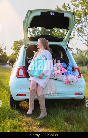 Jeune femme tenant un élégant étui de pique-nique turquoise à côté d'un coffre de voiture rempli de fleurs pendant l'heure dorée dans la campagne, escapade estivale relaxante Banque D'Images