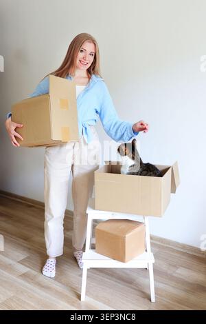 Femme souriante portant une boîte de déménagement tandis que son chat se détend à l'intérieur d'une autre boîte dans un appartement moderne lumineux. Banque D'Images