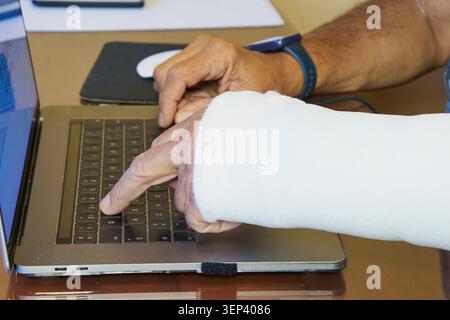 Homme avec une main blessée dans un plâtre travaillant sur un ordinateur portable, surmontant une blessure au doigt Banque D'Images
