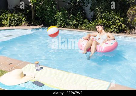 Femme asiatique flottant sur le tube de beignet gonflable rose dans la piscine avec ballon de plage rebondissant, espace de copie Banque D'Images