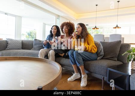Diverses amies féminines jouant à un jeu de console sur un canapé tenant des contrôleurs et mangeant du bol Banque D'Images